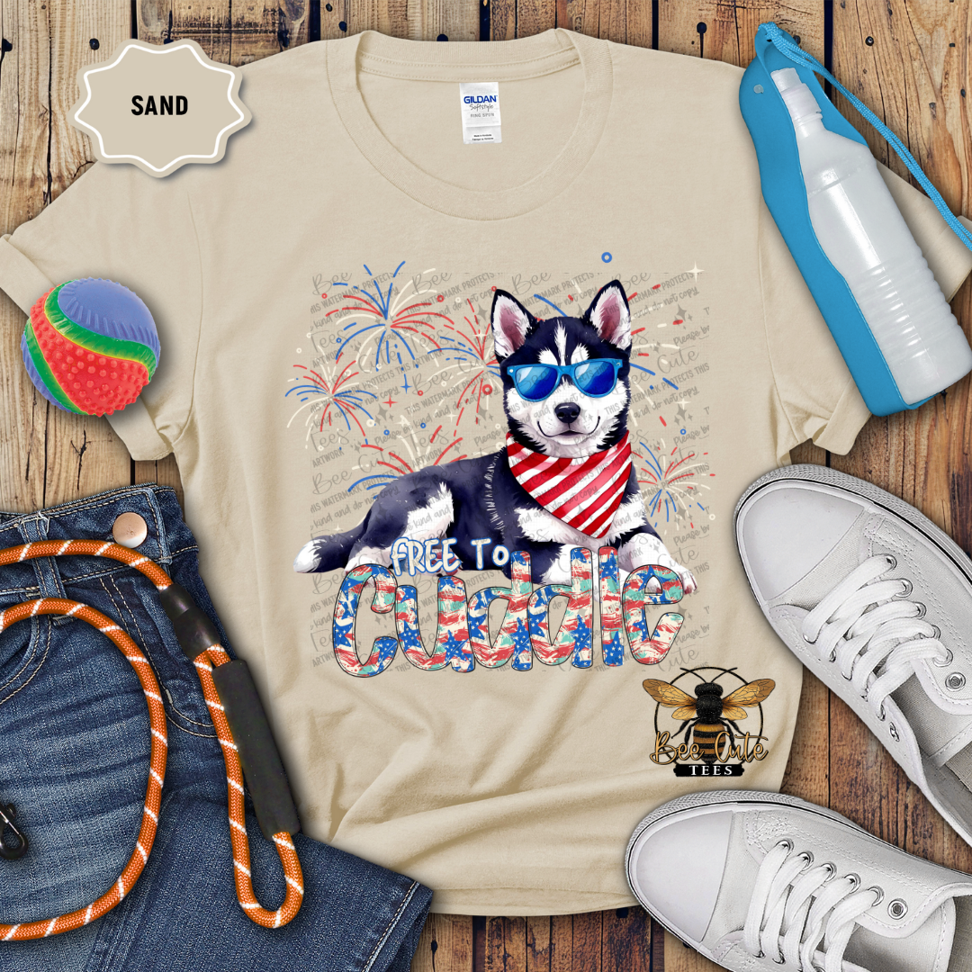 A beige colored t-shirt with a graphic print of a husky dog wearing sunglasses and the text 'Free To Cuddle', along with Fourth of July-themed elements such as fireworks. The shirt is displayed with a pair of jeans, a pair of white sneakers, a blue water bottle, and a beach ball.