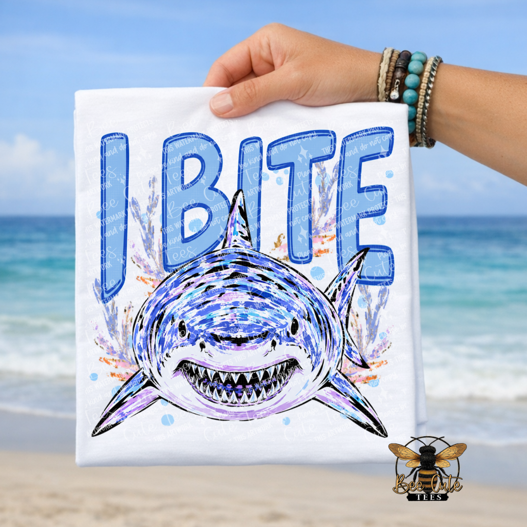 A hand holds a white T-shirt featuring a blue and purple shark graphic with the text 'I BITE' in large blue letters, set against a beach background with ocean and sand visible.