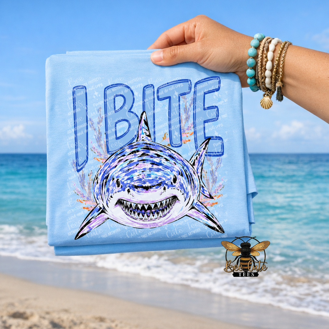 A folded light blue T-shirt with a graphic of a shark and the text 'I BITE' is held up by a person's hand against a beach background. The shirt also features a small bee logo near the bottom right.
