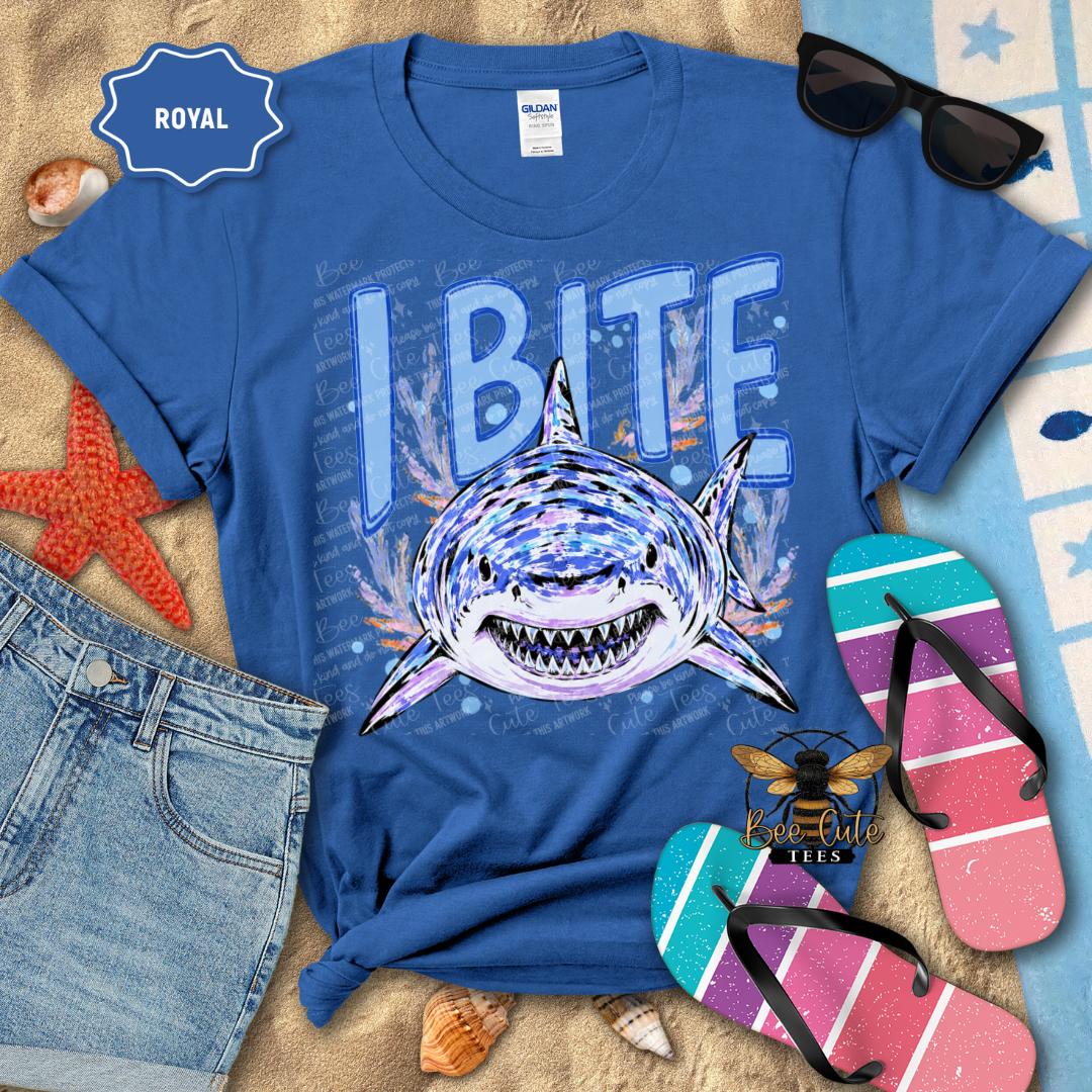 A royal blue graphic t-shirt featuring a shark and the text 'I BITE' in a beach-themed flat lay with sunglasses, denim shorts, flip-flops, and seashells.