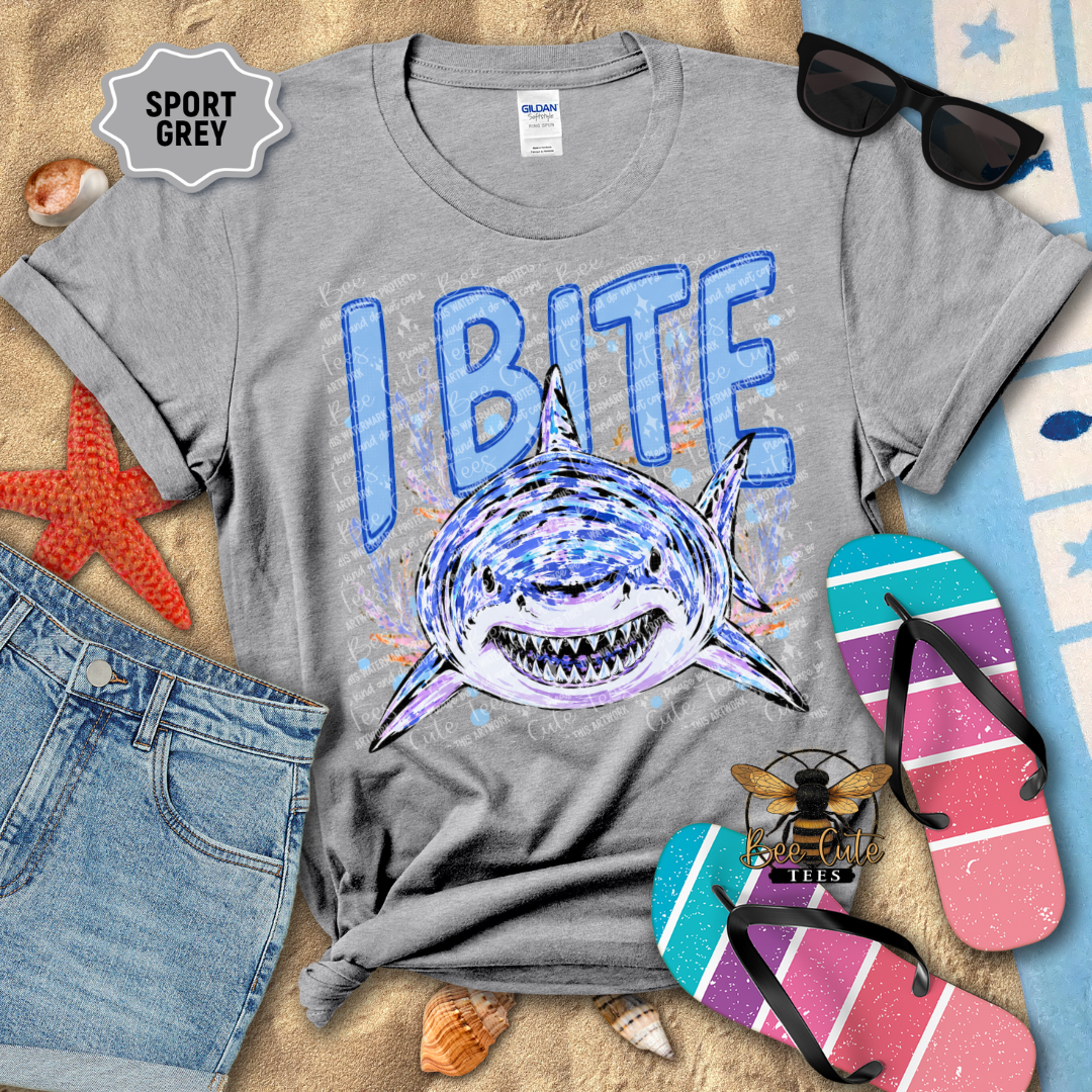 A heather grey t-shirt featuring a graphic of a shark biting a piece of paper with the text 'I BITE'. The shirt is styled in a flat-lay arrangement on a sandy beach-themed background with sunglasses, flip-flops, a denim shorts, and starfish.