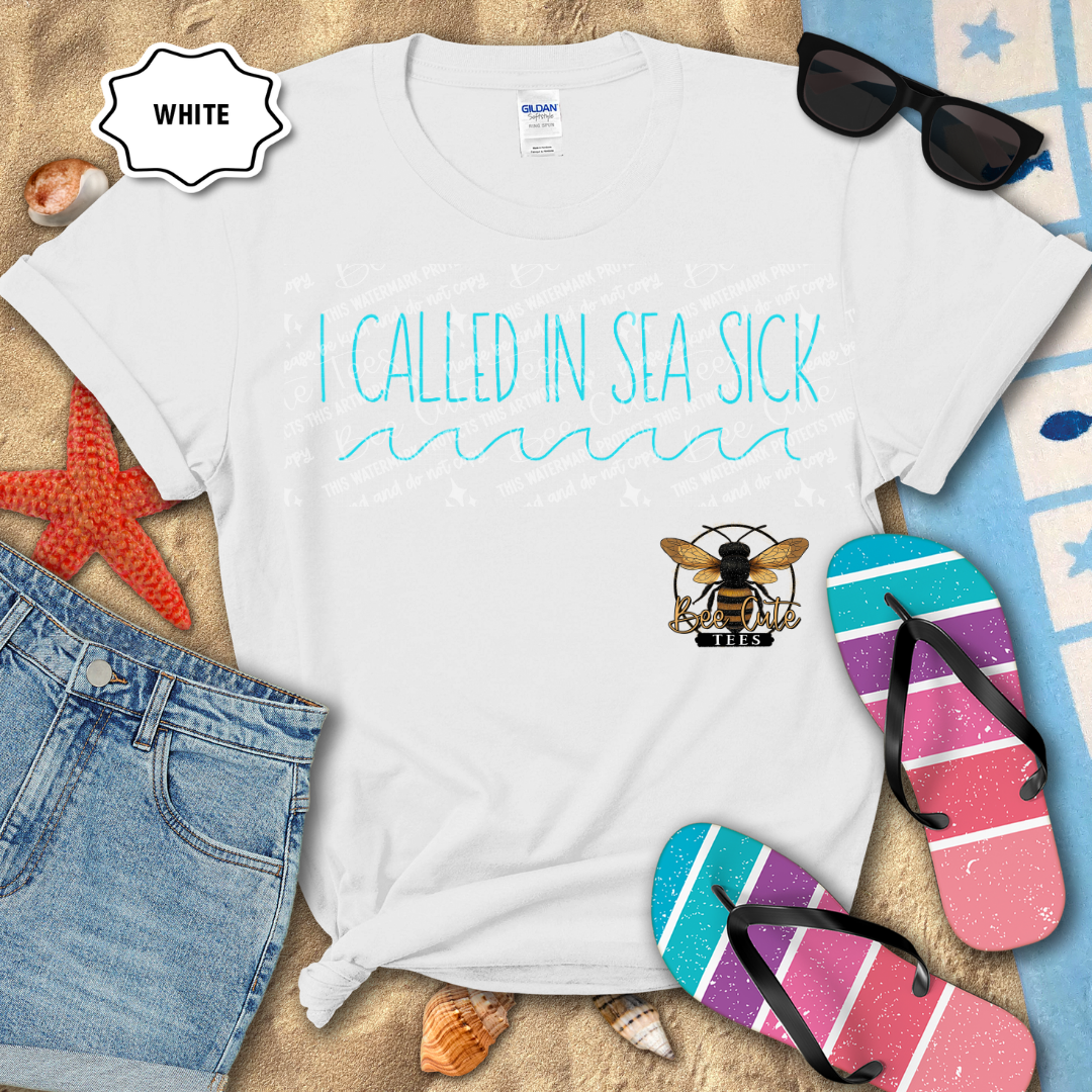 White t-shirt with 'I Called In Sea Sick' text and bee logo, surrounded by beach items on sand.