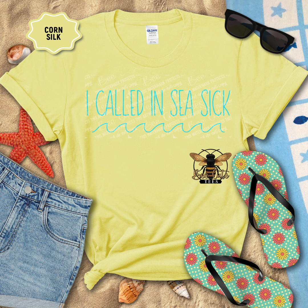 Yellow t-shirt with 'I Called In Sea Sick' text and bee logo, surrounded by beach accessories on sand.