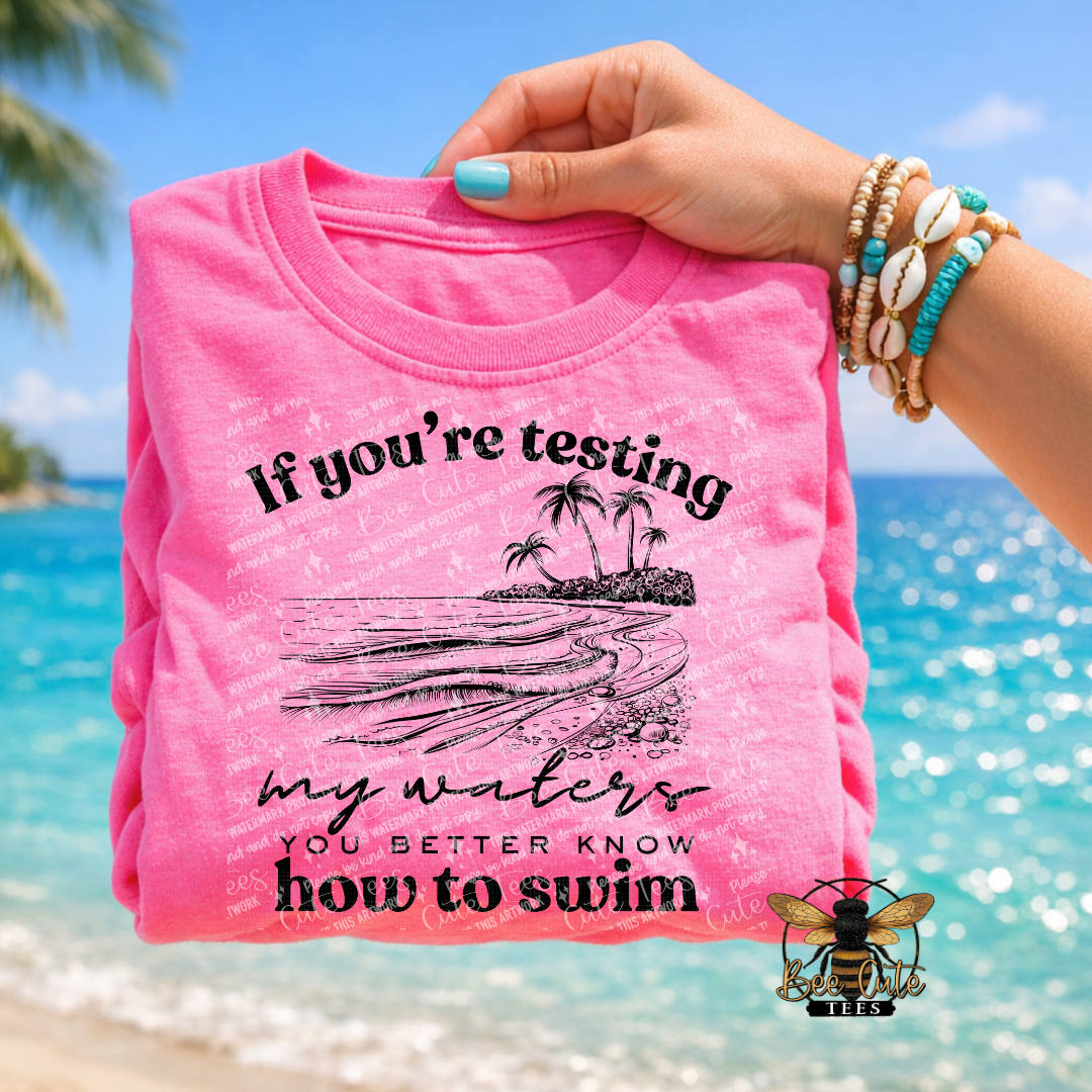 Pink t-shirt with beach graphic and text held by a hand against a beach background