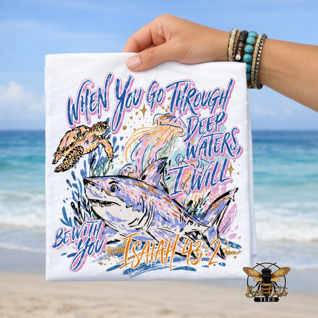 A hand holds a white graphic T-shirt against a beach background. The shirt features a colorful illustration of a shark, a turtle, and a fish with the text 'WHEN YOU GO THROUGH DEEP WATERS I WILL BE WITH YOU ISAIAH 43:2'.