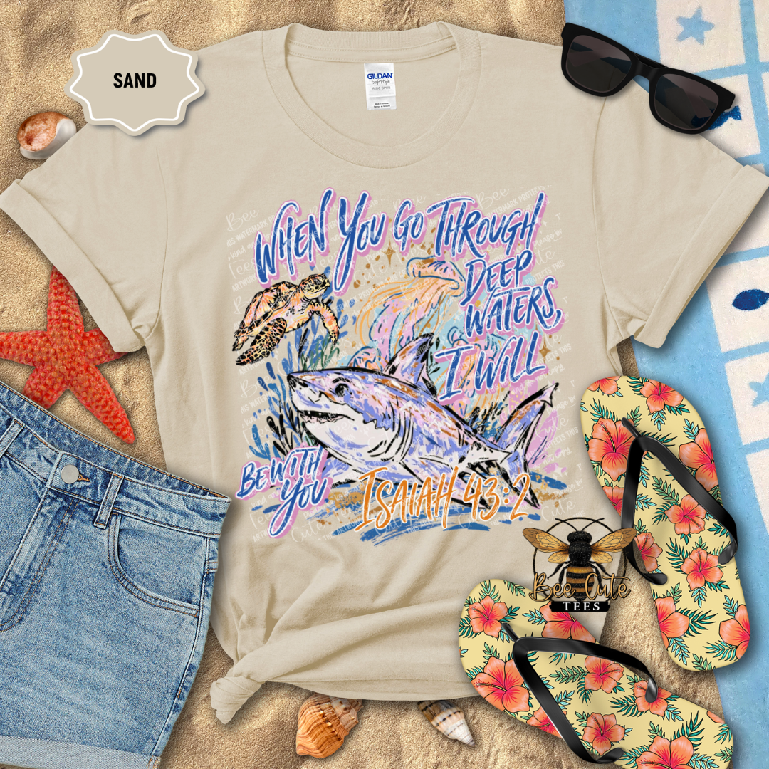 A beige graphic t-shirt laid flat on a sandy surface with beach-themed props including sunglasses, flip-flops, denim shorts, shells, and a star-patterned cloth. The shirt graphic shows a shark, turtle, and colorful text reading 'When You Go Through Deep Waters, I Will Be With You Isaiah 43:2'.