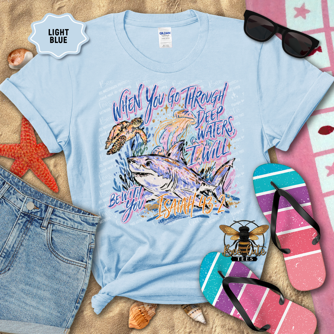 A light blue graphic t-shirt laid flat on a sandy surface with beach-themed props including sunglasses, flip-flops, denim shorts, shells, and a starfish. The shirt features a shark and turtle illustration with the text 'When You Go Through Deep Waters Thou Wilt Be with You Est.