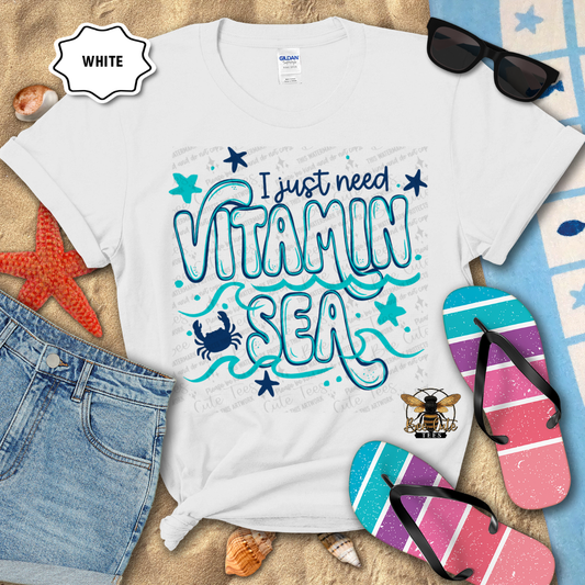 White t-shirt with 'I just need Vitamin Sea' design, sunglasses, denim shorts, and flip-flops on a beach-themed background.