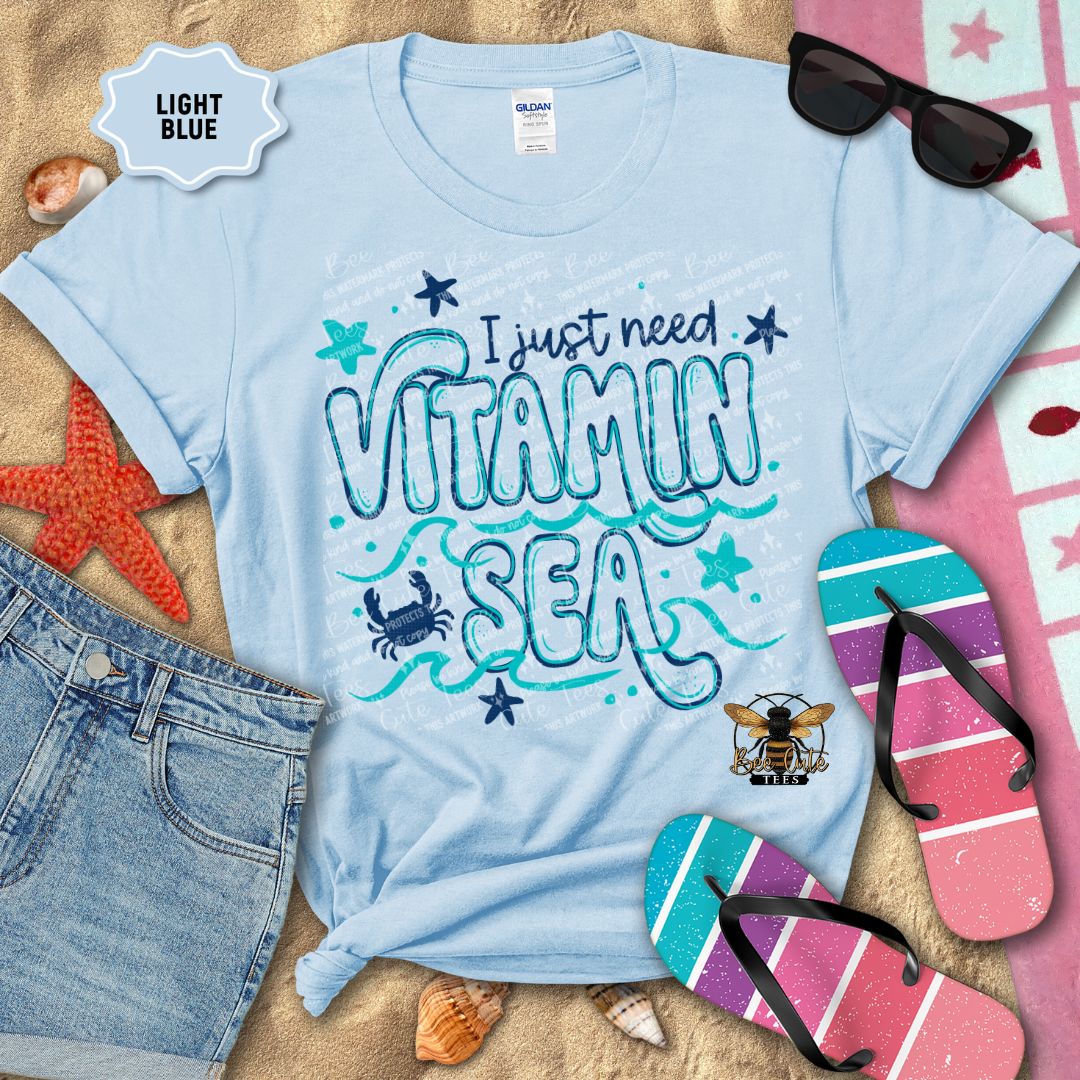 Light blue t-shirt with 'I just need vitamin sea' design on a sandy background with beach accessories.