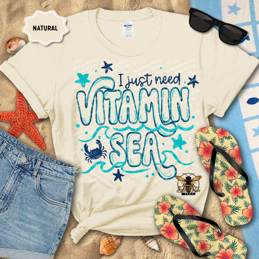 Beige t-shirt with 'I just need Vitamin Sea' design on a beach-themed background with sunglasses, flip-flops, and starfish.