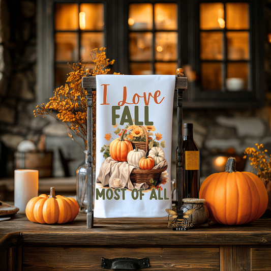 A white tea towel with a fall harvest design and the text 'I Love FALL MOST OF ALL' displayed on a wooden stand on a rustic wooden table, surrounded by pumpkins, candles, and autumn decorations.