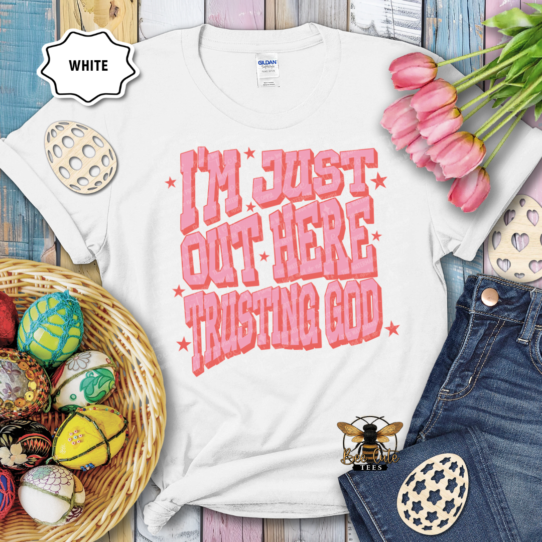 White t-shirt with pink text 'I'm just out here trusting God' on a wooden surface with Easter decorations.