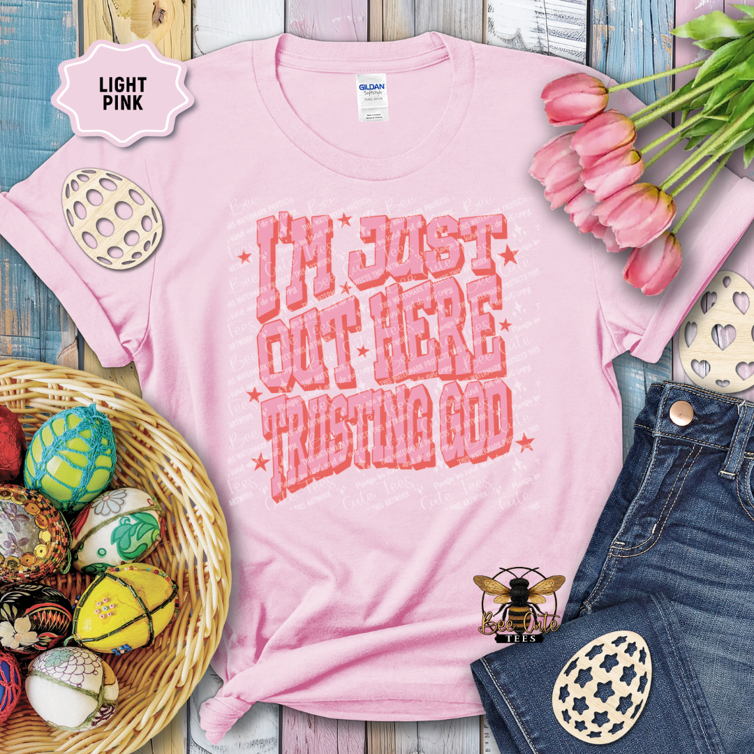 Light pink t-shirt with 'I'm just out here trusting God' text, surrounded by Easter decorations on a wooden surface.