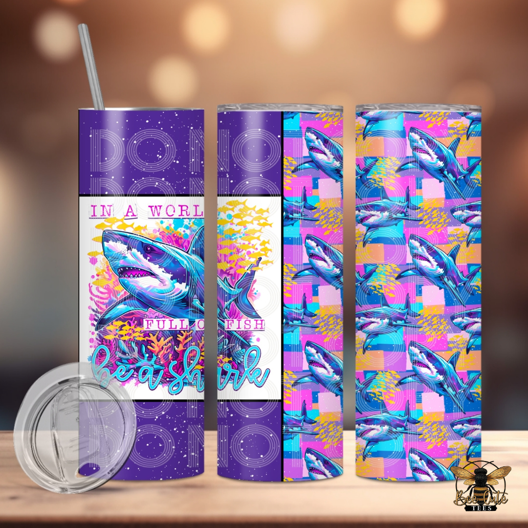 Colorful 20 oz tumbler featuring a vibrant shark design with coral reef accents and “In a World Full of Fish, Be a Shark” wording, paired with a bold shark pattern wrap in purple, pink, and turquoise tones, displayed with a lid and metal straw against a softly lit background.
