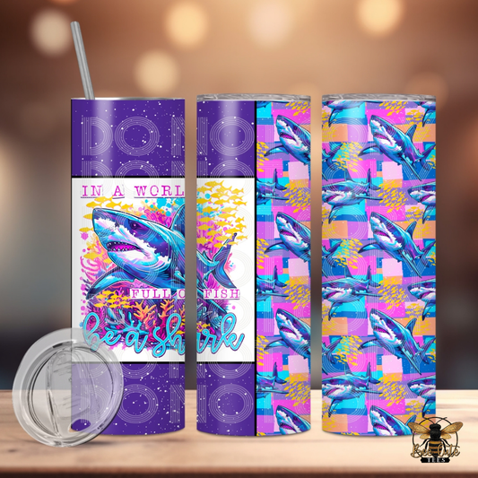 Colorful 20 oz tumbler featuring a vibrant shark design with coral reef accents and “In a World Full of Fish, Be a Shark” wording, paired with a bold shark pattern wrap in purple, pink, and turquoise tones, displayed with a lid and metal straw against a softly lit background.
