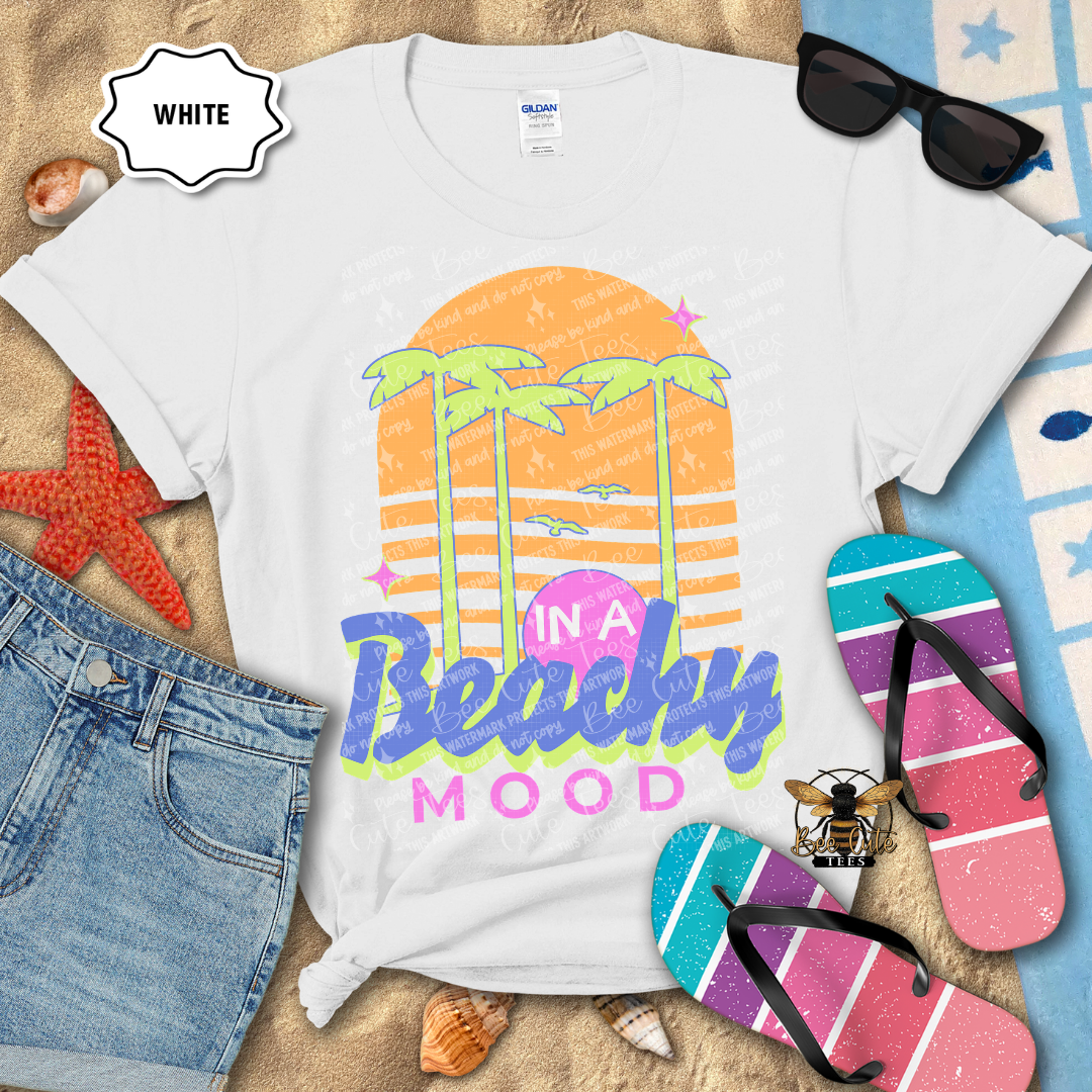 White t-shirt with 'In a Beachy Mood' design, sunglasses, flip-flops, and beach accessories on a sandy background.