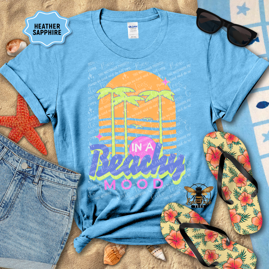 Blue t-shirt with 'In a Beachy Mood' design, sunglasses, flip-flops, and beach accessories on sand.