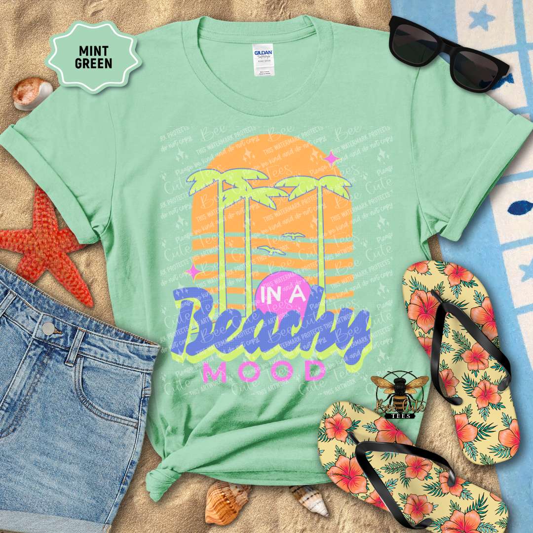 Mint green t-shirt with 'In a Beachy Mood' design, sunglasses, flip-flops, and beach accessories on a sandy background.