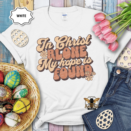 White t-shirt with 'In Christ Alone My Hope is Found' text, surrounded by Easter decorations on a wooden surface.