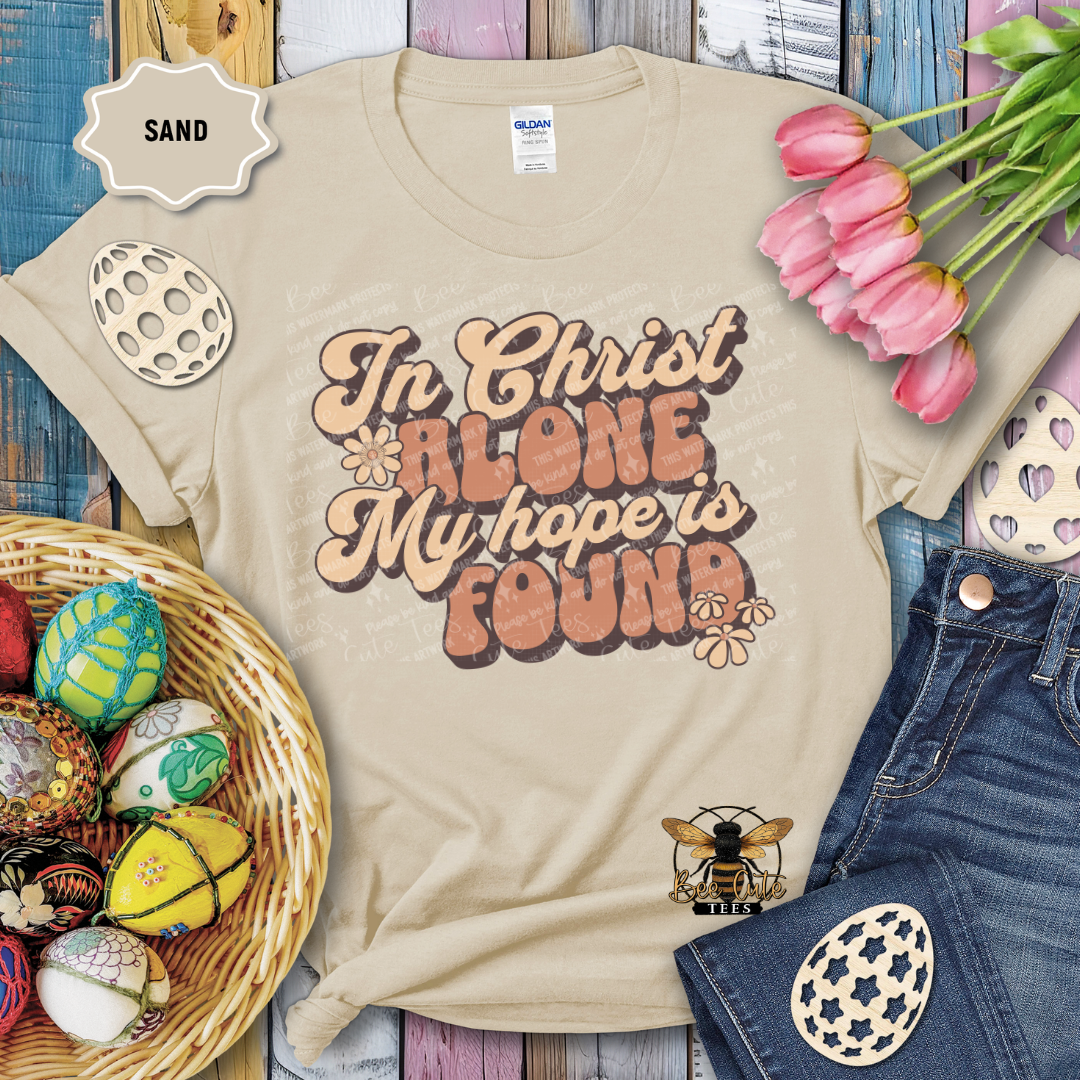 Beige t-shirt with 'In Christ Alone My hope is Found' text, surrounded by Easter decorations on a wooden surface.