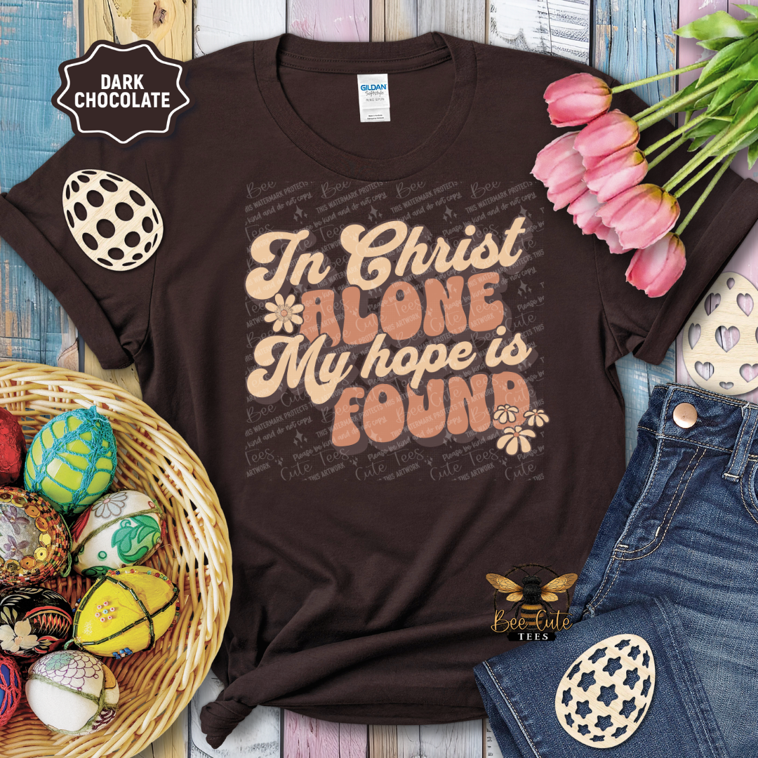 Brown t-shirt with 'In Christ Alone My hope is Found' text, surrounded by Easter decorations on a wooden surface.