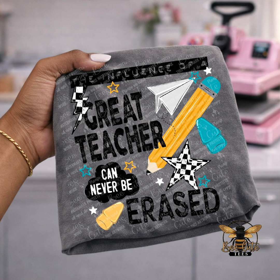 A hand holds a dark gray t-shirt featuring a school-themed graphic with the text 'GREAT TEACHER CAN NEVER BE ERASED' and illustrations of a yellow pencil, blue eraser, paper airplane, and stars. The background is a blurred pink and gray craft room setting.
