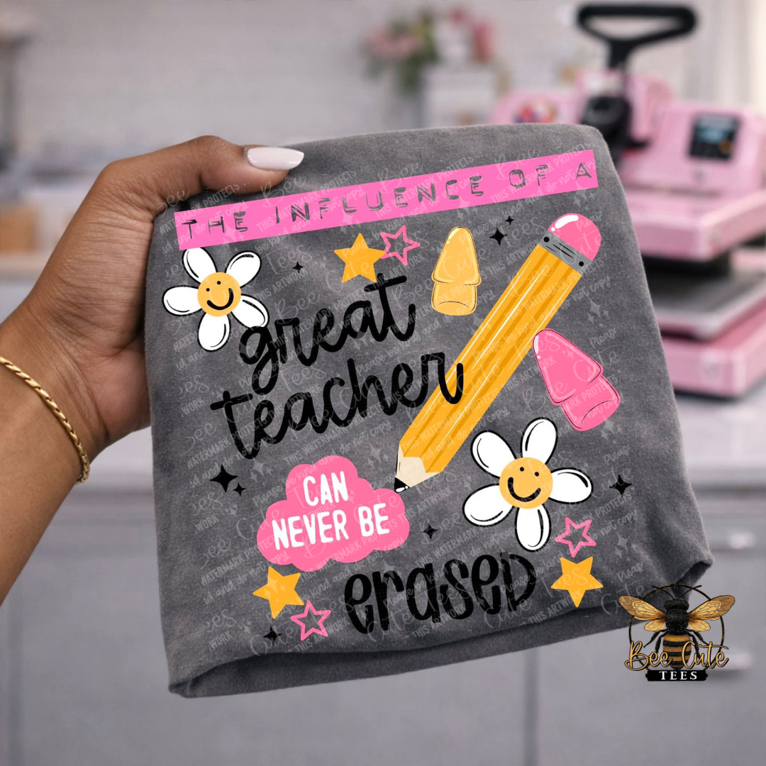 A hand holds a dark gray t-shirt featuring a colorful graphic design that reads 'the influence of a great teacher can never be erased' with a yellow pencil, smiley daisies, stars, and pink clouds.