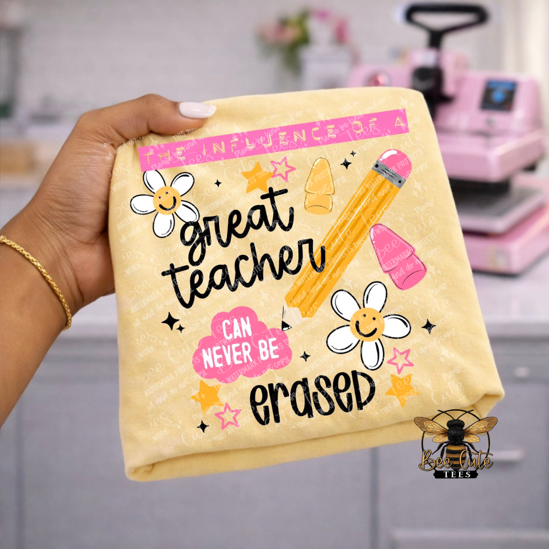 A hand holds a folded light yellow t-shirt featuring a 'great teacher can never be erased' graphic with a pencil, flowers, and stars. The background shows a blurred pink heat press machine in a craft room setting.