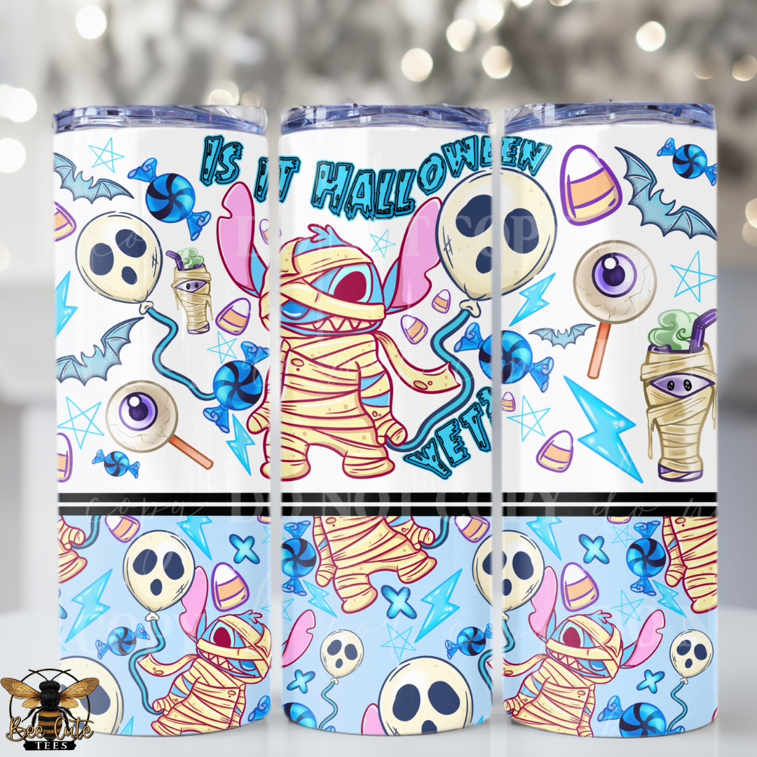 Three tall insulated tumblers with clear lids are shown on a softly blurred festive background. The tumblers have a Halloween-themed wrap design featuring cartoon mummies, skulls, bats, candy corn, eyeballs, lightning bolts, and the text 'IT IS HALLOWEEN YET'.