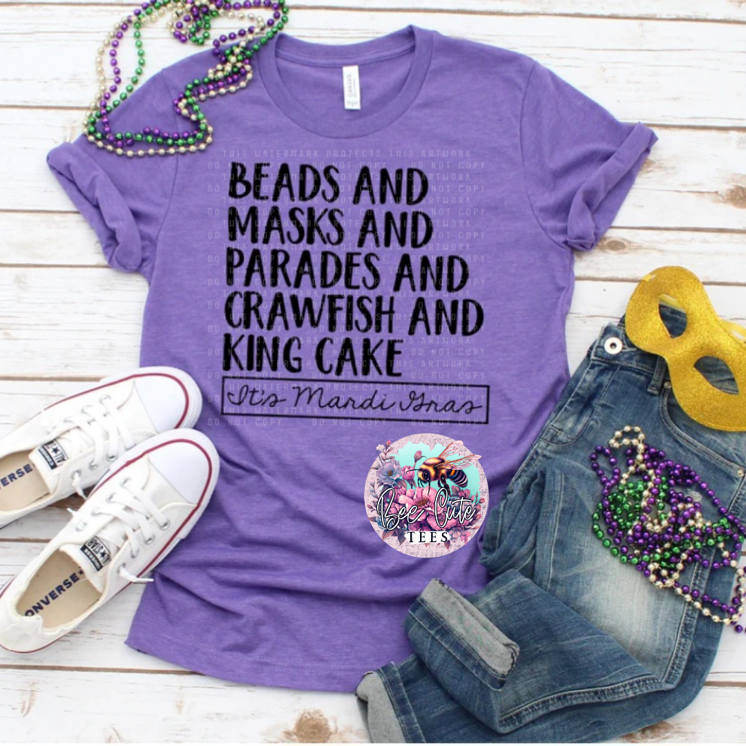 A purple t-shirt with text 'BEADS AND MASKS AND PARADES AND CRAWFISH AND KING CAKE' by Mandi Jardin, displayed on a wooden background with a pair of blue jeans, a pair of white sneakers, a mask, and beaded necklaces.