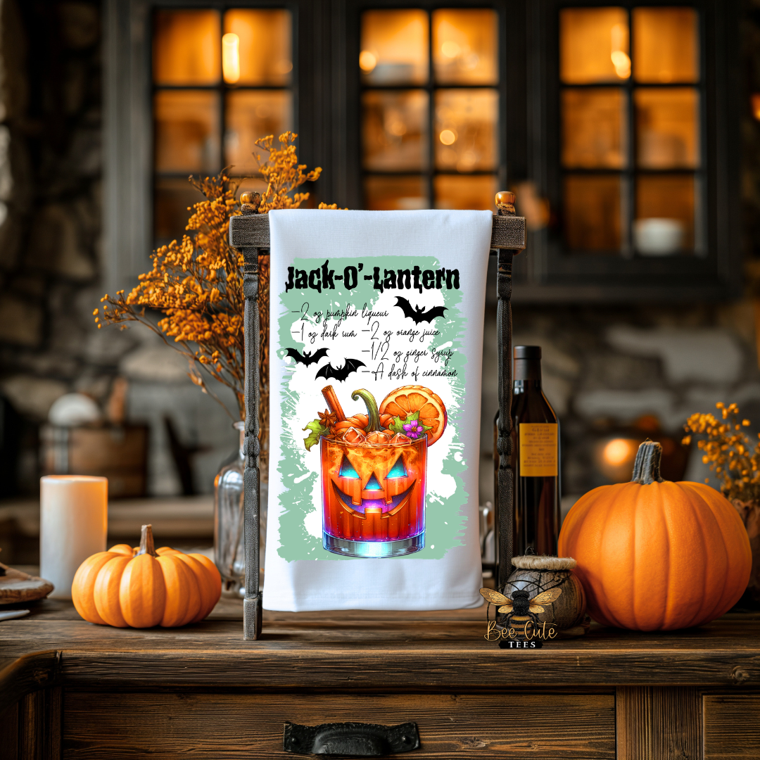 A white kitchen tea towel with a Halloween-themed 'Jack-O-Lantern' cocktail print is displayed on a wooden stand on a rustic table, surrounded by pumpkins, candles, and autumn decorations.