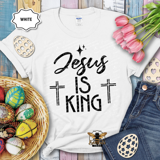 White t-shirt with 'Jesus is King' text, surrounded by Easter decorations on a wooden surface.