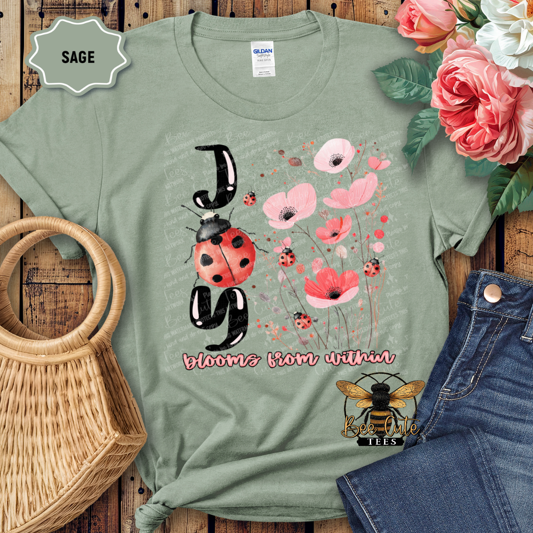 Sage green t-shirt with floral and ladybug design on a wooden surface with flowers and jeans.