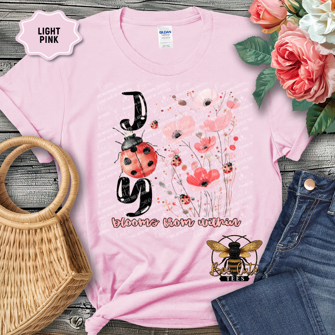 Light pink t-shirt with ladybug and bee logo, surrounded by flowers and denim jeans on a wooden surface.