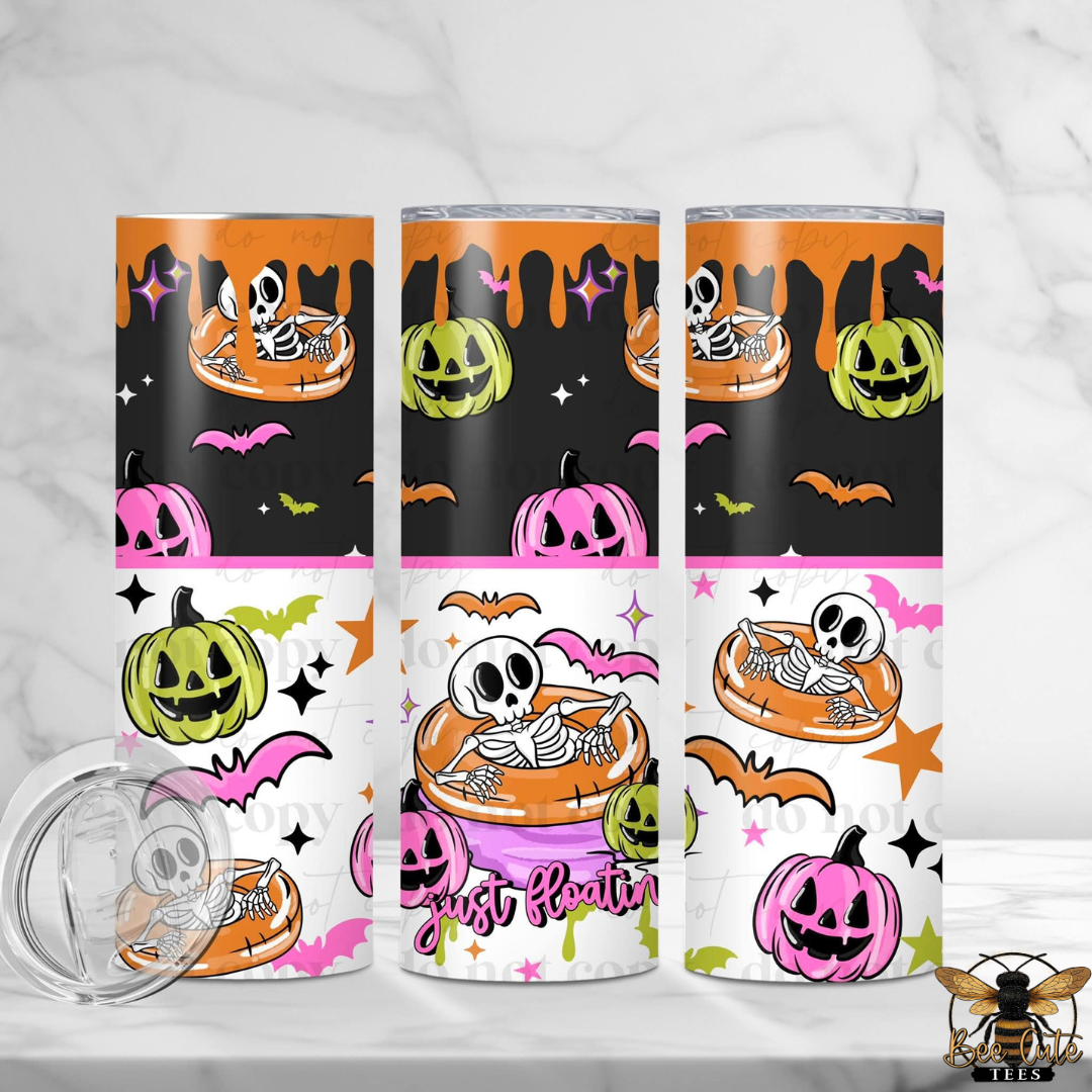 Three tall skinny tumblers with a Halloween-themed graphic print featuring skeletons in orange floaties, jack-o'-lanterns, bats, and stars on black and white backgrounds. A clear plastic lid is shown in the foreground on a light marble surface.