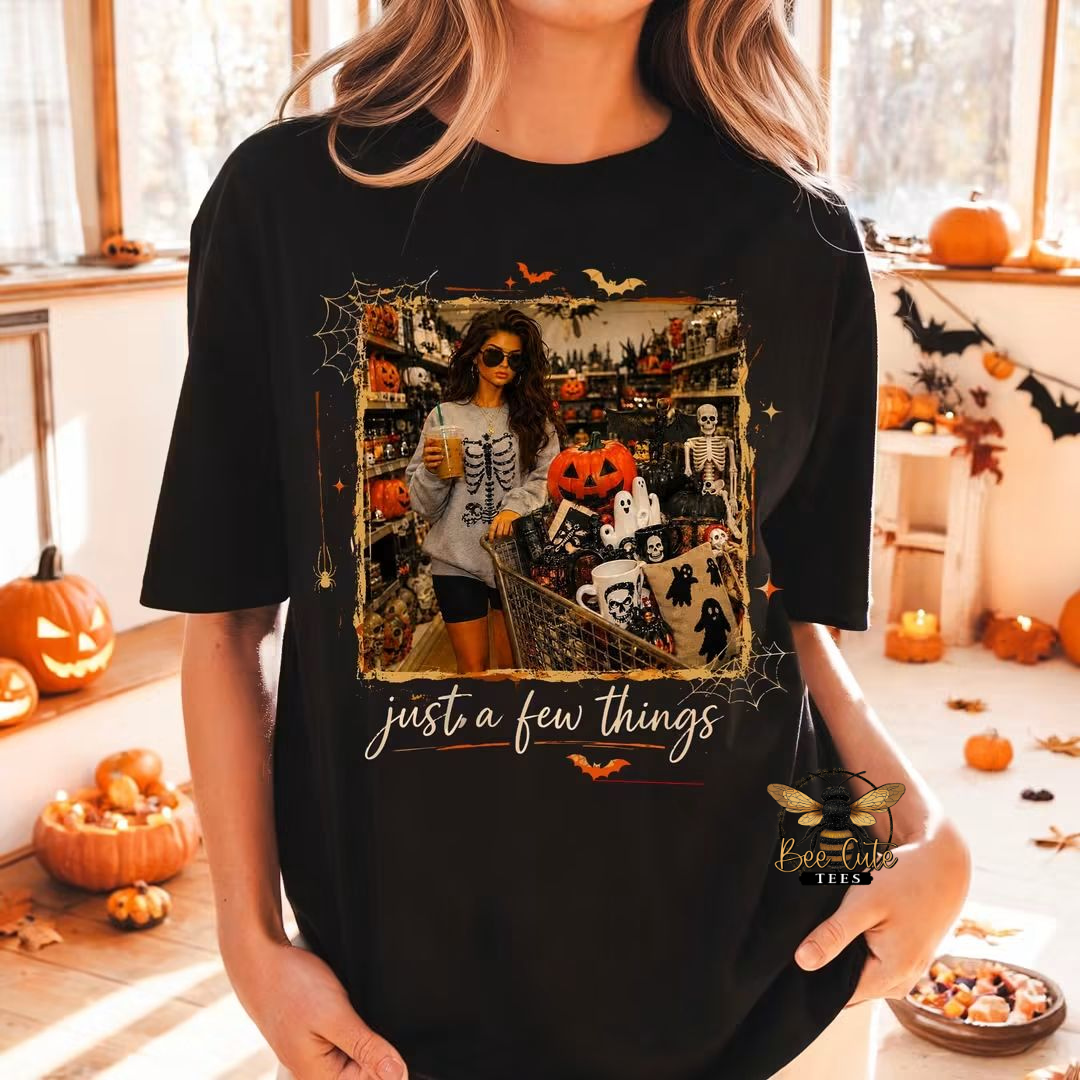 Black Halloween graphic tee featuring a vintage-style scene of a woman shopping for spooky décor with a cart full of pumpkins, ghosts, skeletons, and Halloween finds, framed with bats and spiderwebs and the phrase “just a few things.”