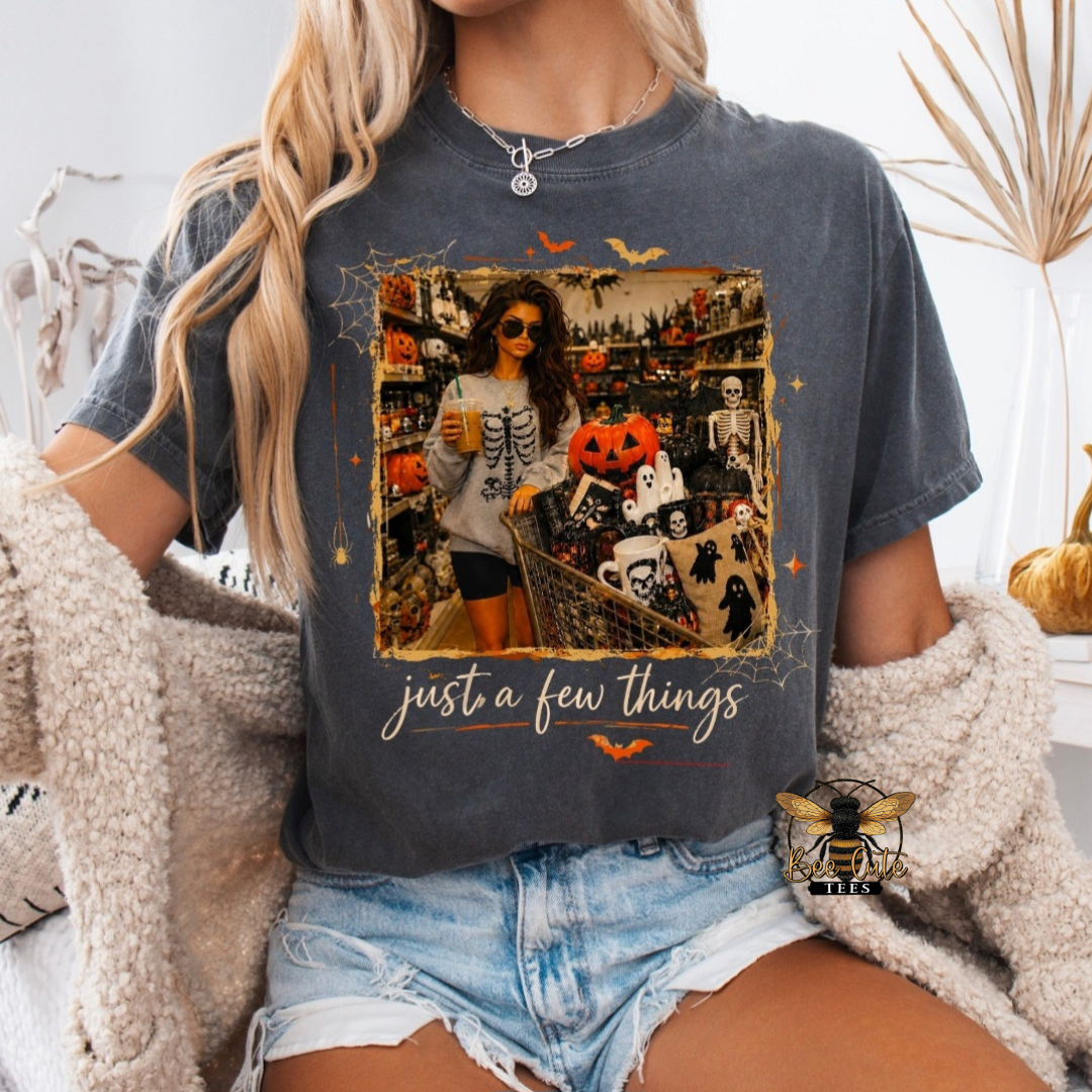 Charcoal gray Halloween graphic tee featuring a spooky shopping scene with a woman and a cart filled with pumpkins, ghosts, skeletons, mugs, and Halloween décor, accented with bats and spiderwebs and the phrase “just a few things.”