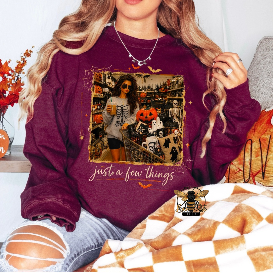 Maroon Halloween sweatshirt featuring a vintage-style spooky shopping scene with a woman and a cart overflowing with pumpkins, ghosts, skeletons, and Halloween décor, framed by bats and spiderwebs with the phrase “just a few things.”