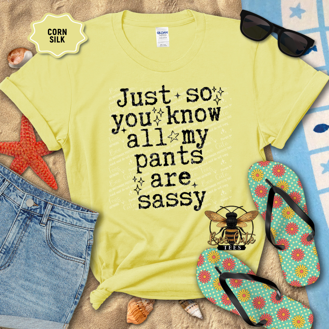 Yellow t-shirt with 'Just so you know all my pants are sassy' text, surrounded by beach items on sand.