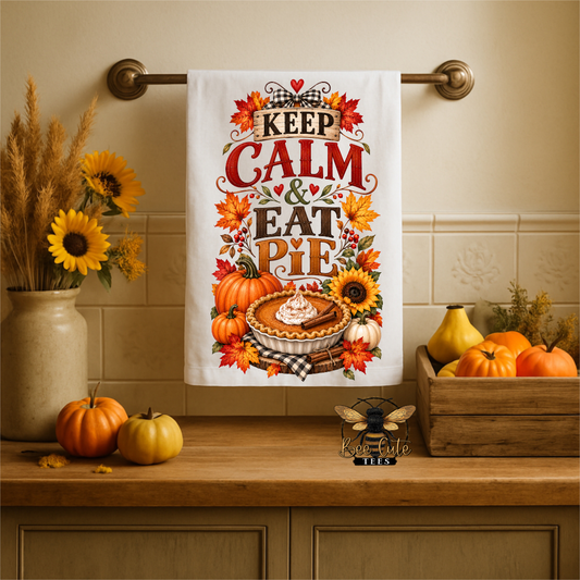 A white kitchen towel with a colorful autumn 'Keep Calm & Eat Pie' graphic hangs on a brass towel bar over a wooden counter. The scene is styled with fall decorations including pumpkins, sunflowers, and a wooden crate.