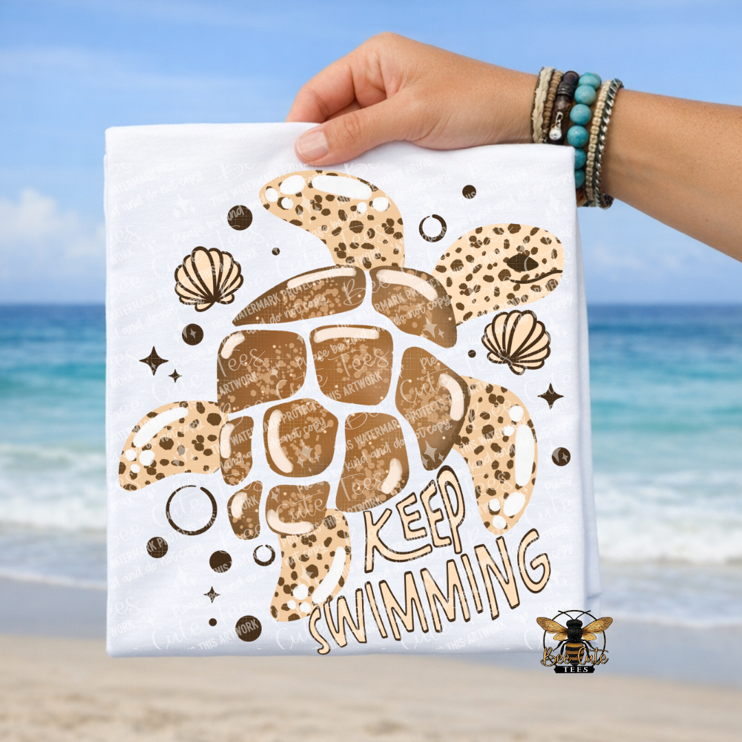White T-shirt with turtle design and 'Keep Swimming' text held by a hand on a beach.