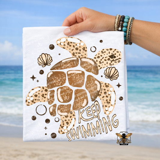 White T-shirt with turtle design and 'Keep Swimming' text held by a hand on a beach.