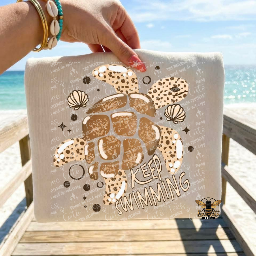 Beige t-shirt with turtle design and 'Keep Swimming' text held by a hand on a beach.