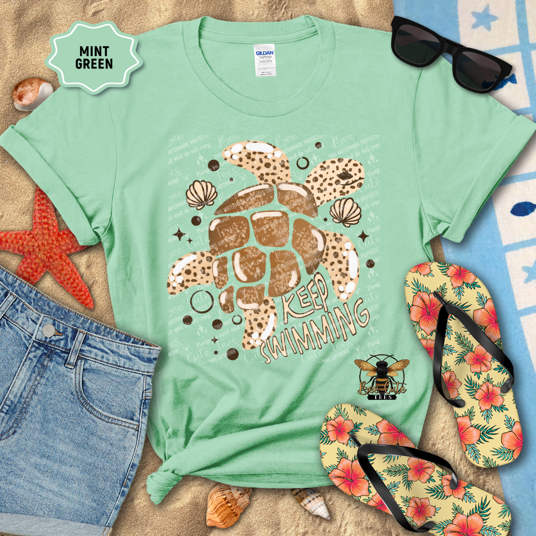 Mint green t-shirt with a turtle graphic and text, surrounded by beach accessories on a sandy background.