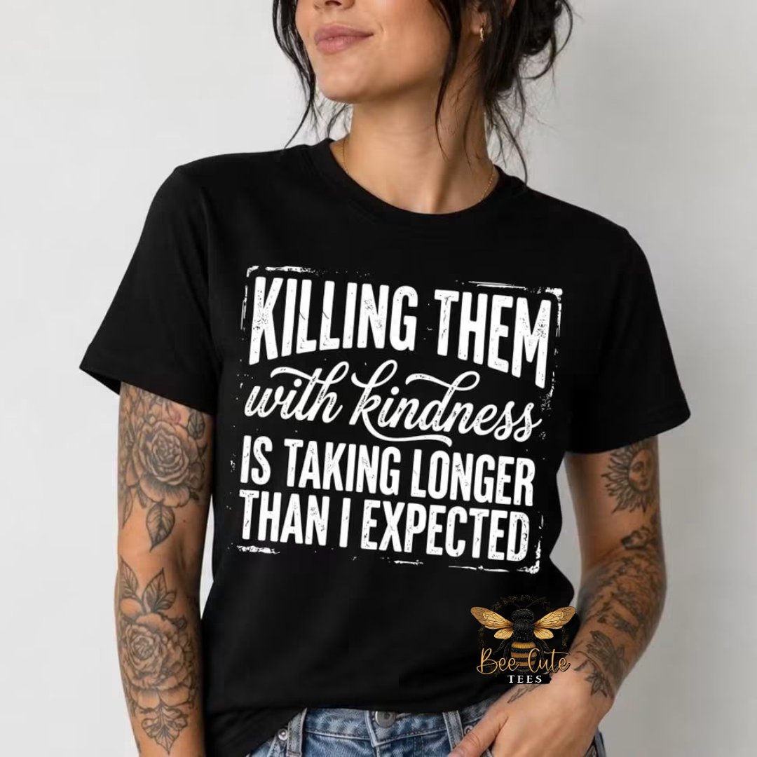 A woman wearing a black short-sleeve graphic T-shirt with distressed white text that reads, 'KILLING THEM with kindness IS TAKING LONGER THAN I EXPECTED.' She has visible tattoos on both arms and is wearing blue jeans against a plain light gray background.