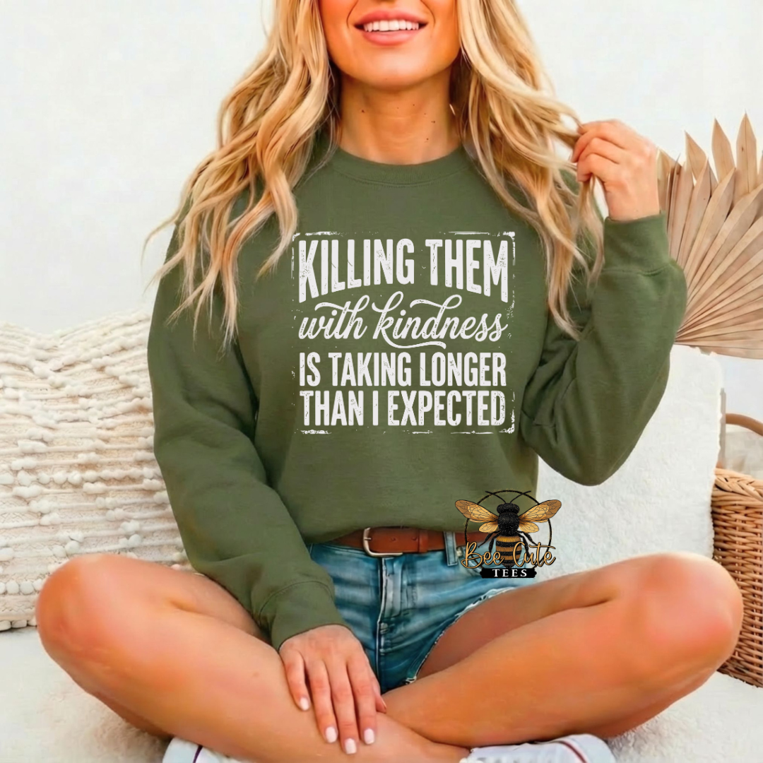 A woman sitting cross-legged on a white couch wearing an olive green crewneck sweatshirt with the text 'KILLING THEM with kindness IS TAKING LONGER THAN I EXPECTED' in white. She is wearing blue denim shorts, and a decorative dried palm leaf and wicker basket are visible in the background.
