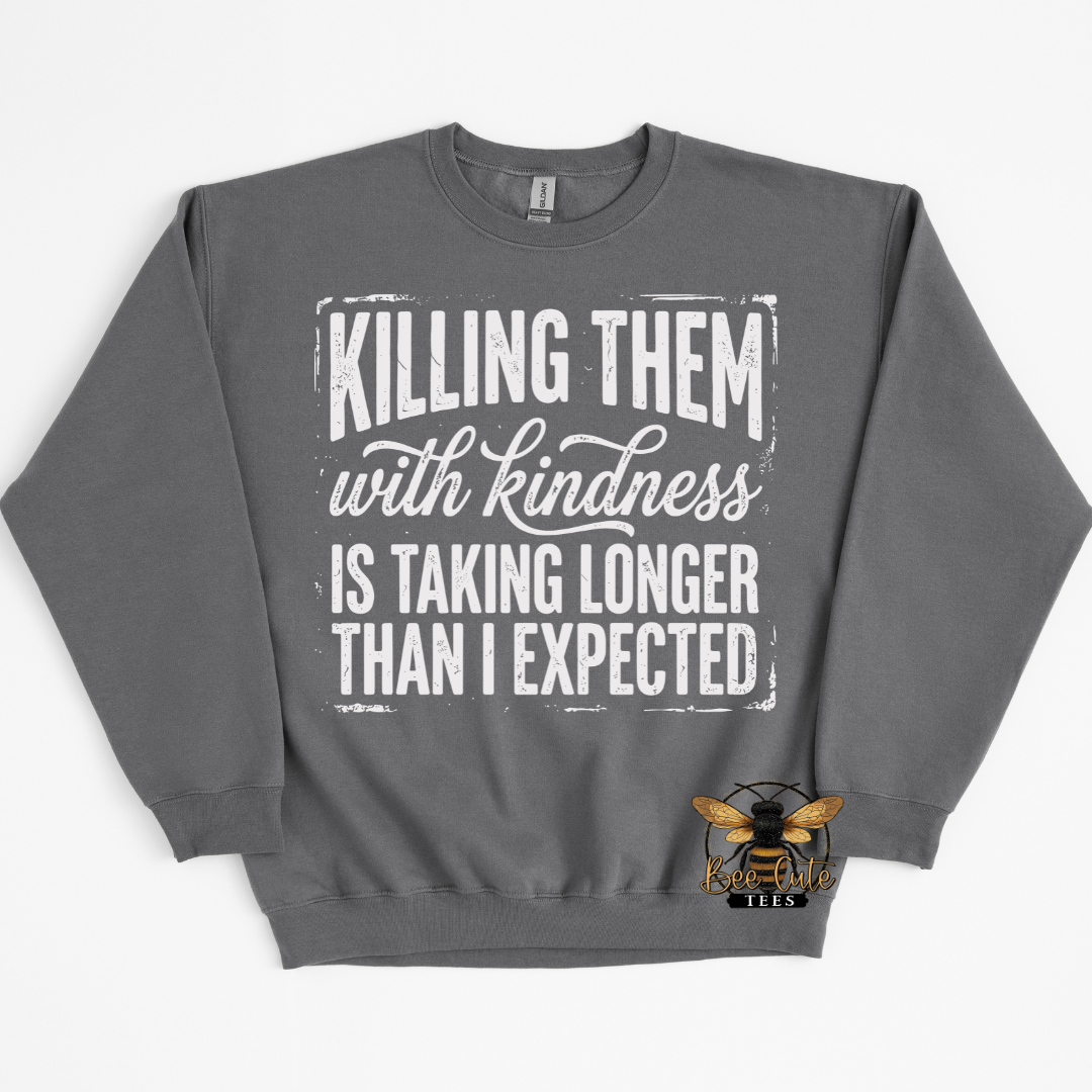 A dark gray crewneck sweatshirt with a distressed white graphic text that reads 'KILLING THEM with kindness IS TAKING LONGER THAN I EXPECTED'. A small bee logo for 'Bee Cute Tees' is printed on the bottom right of the sweatshirt.