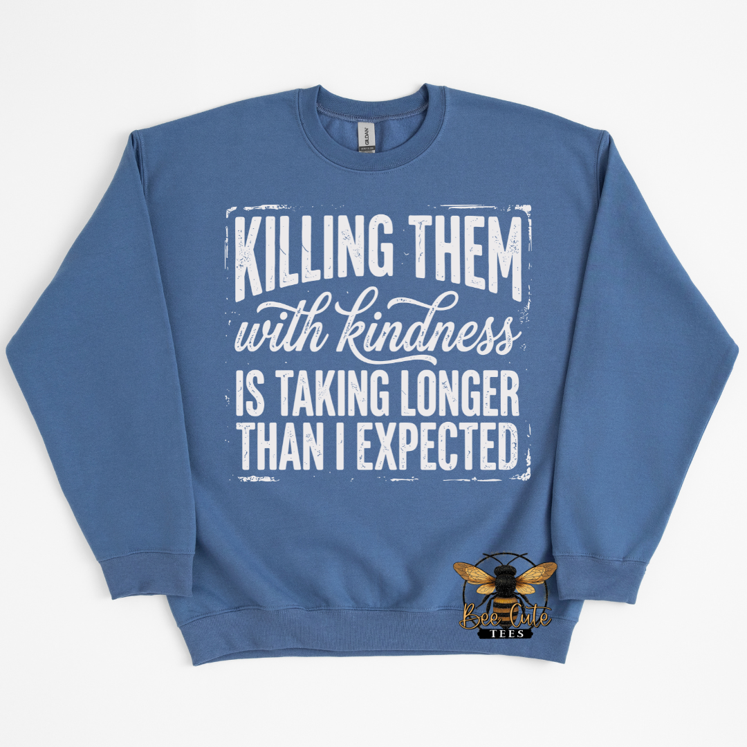 A blue crewneck sweatshirt featuring a white distressed text graphic that reads 'KILLING THEM with kindness IS TAKING LONGER THAN I EXPECTED'. A small bee logo is  near the bottom right of the chest.