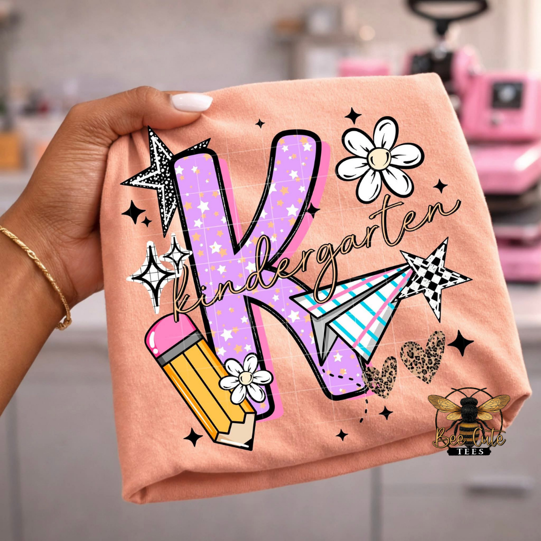 Hand holding an orange t-shirt with 'Kindergarten K' design in a craft room setting