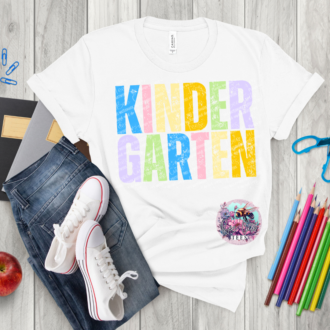 White t-shirt with 'KINDER GARTEN' in colorful letters on a wooden surface with jeans, sneakers, and school supplies.